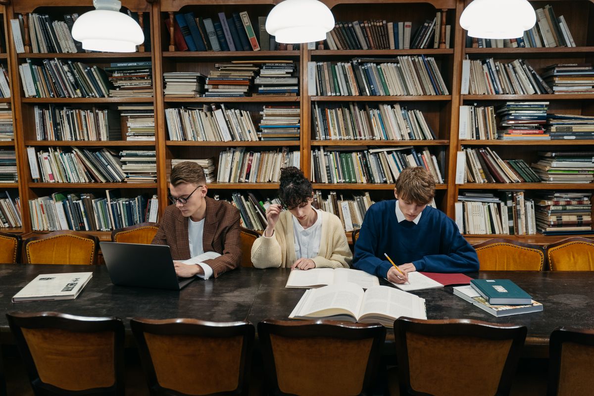 Three students reviewing sources on laptops and books at a library research table