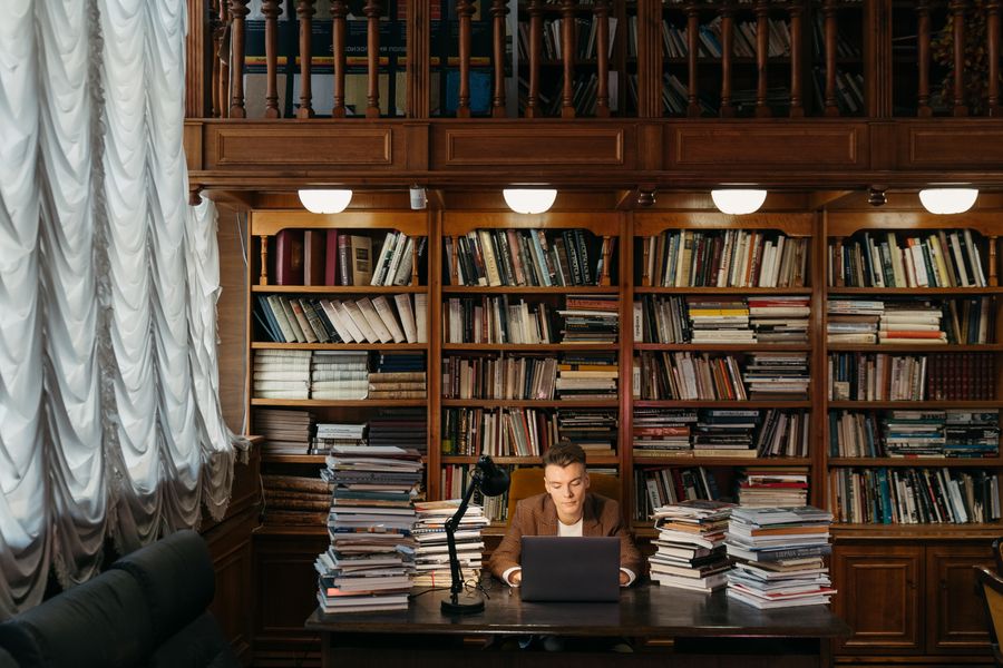A student working on a laptop at a library desk surrounded by stacks of books