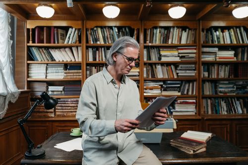 Free Elderly man with eyeglasses reading papers in a library filled with bookshelves. Stock Photo