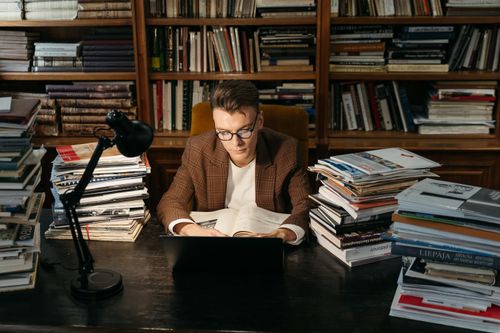 Free A young man intensely studying at a desk surrounded by stacks of books in a library. Stock Photo