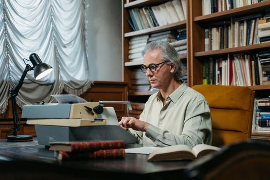 Senior man with glasses and gray hair typing on vintage typewriter in cozy library setting.