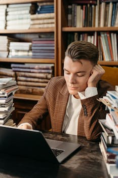 Focused young man sitting at a desk with a laptop, surrounded by books, in a library.