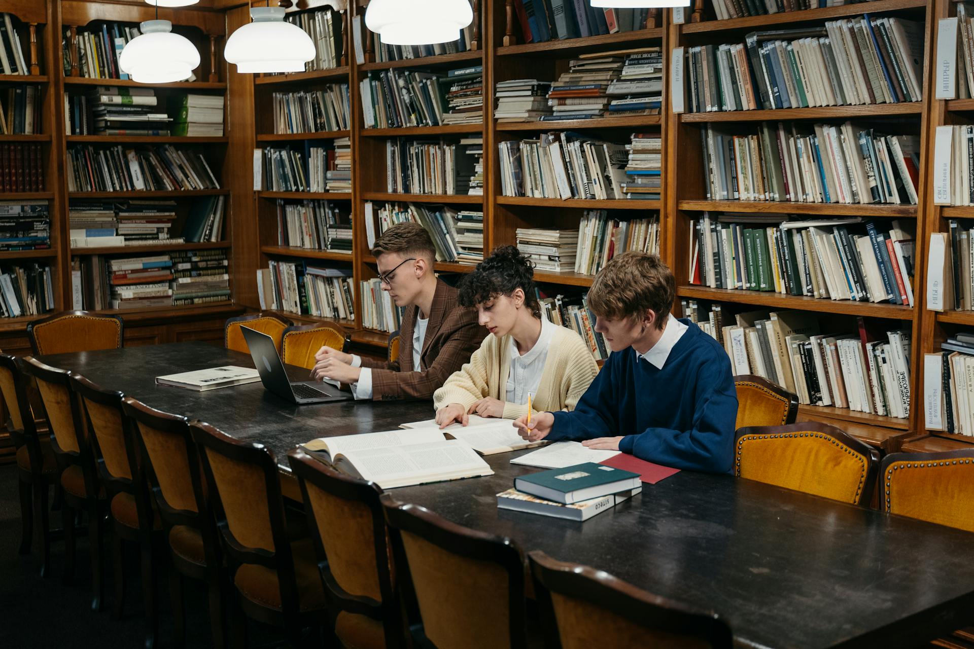 Three students studying and taking notes in a traditional library with wooden bookshelves.