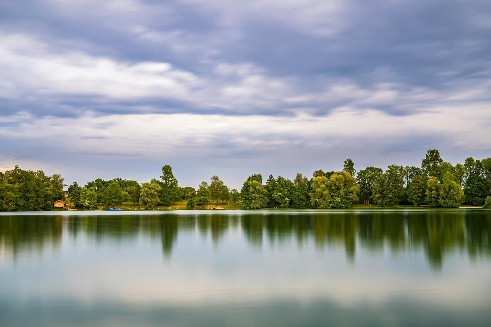 Tranquil lake with tree reflections and a dramatic cloudy sky, captured with long exposure.