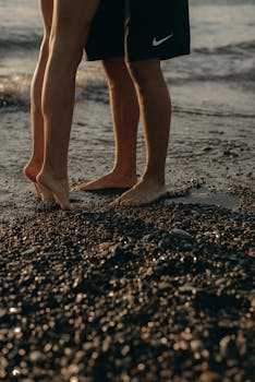 A young couple standing barefoot on a pebble beach at sunset, exuding romance and connection.