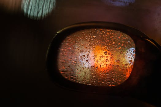 Close-up of raindrops on a car's side mirror with blurred lights at night.