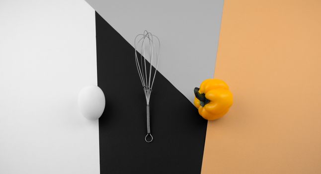 Minimalist flat lay of a whisk, yellow bell pepper, and egg on colorful backgrounds.