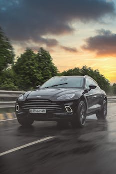 A sleek black Aston Martin SUV driving on a wet highway during a colorful sunset, showcasing luxury and speed.