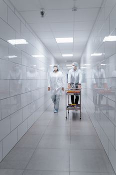 Two healthcare workers in PPE walking with a cart in a sterile hospital hallway.