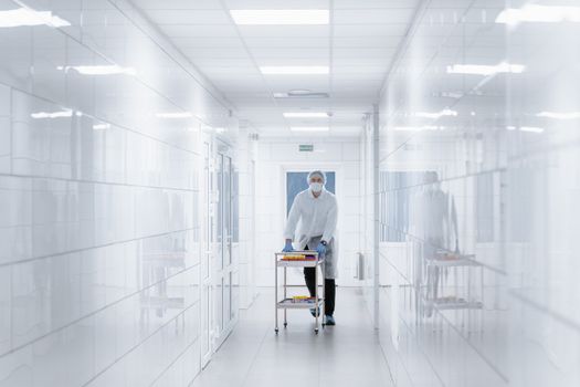 Scientist in protective gear moving samples on a cart through a bright laboratory hallway.