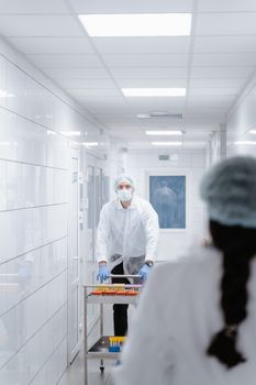 Healthcare professional in PPE pushes a cart with lab samples in a sterile hallway.