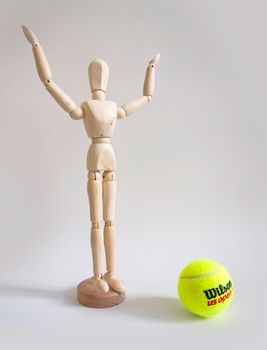 Wooden art mannequin with arms raised beside a US Open tennis ball.