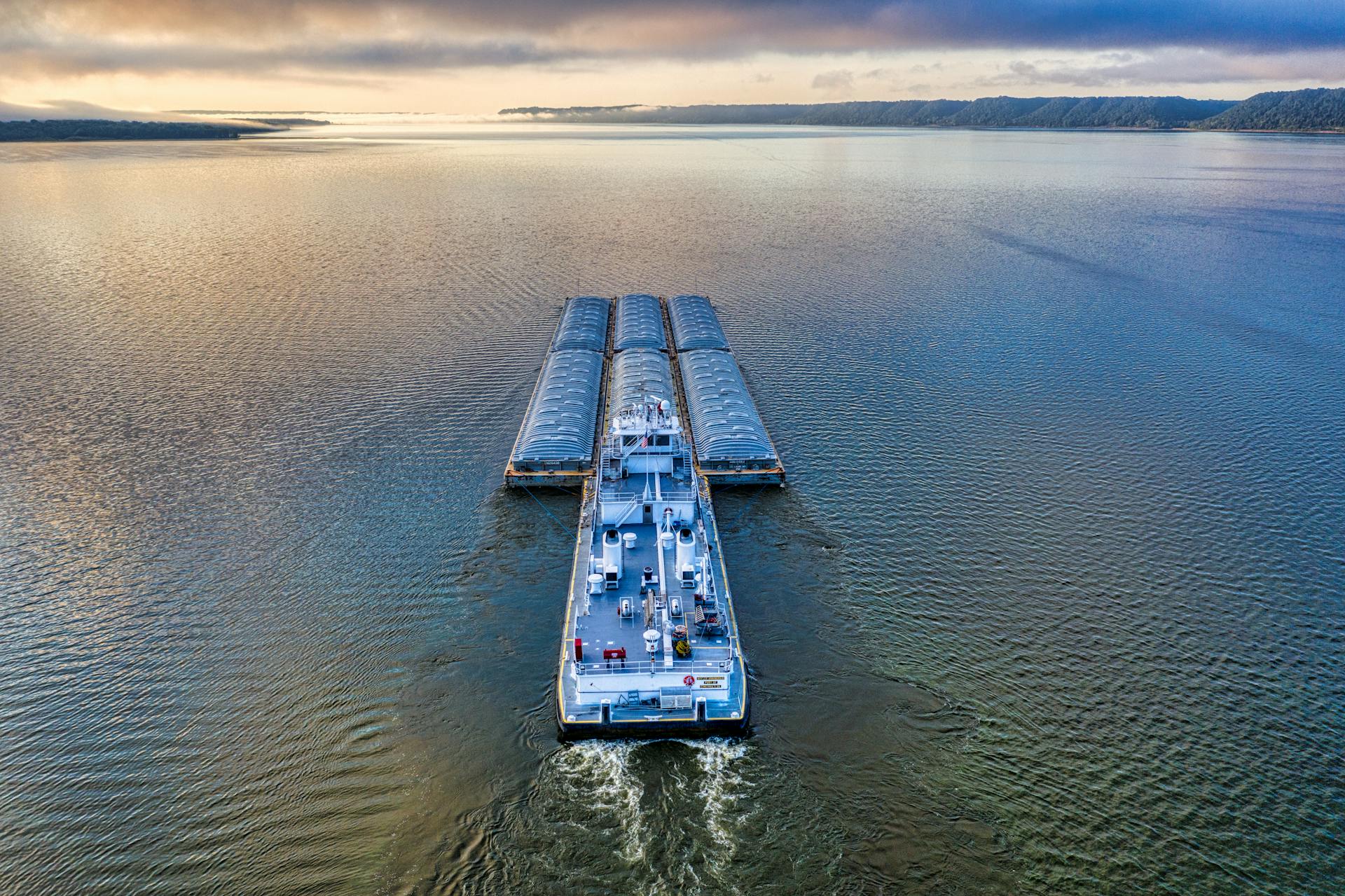 Aerial shot of a barge navigating a vast river under a colorful sunset sky.