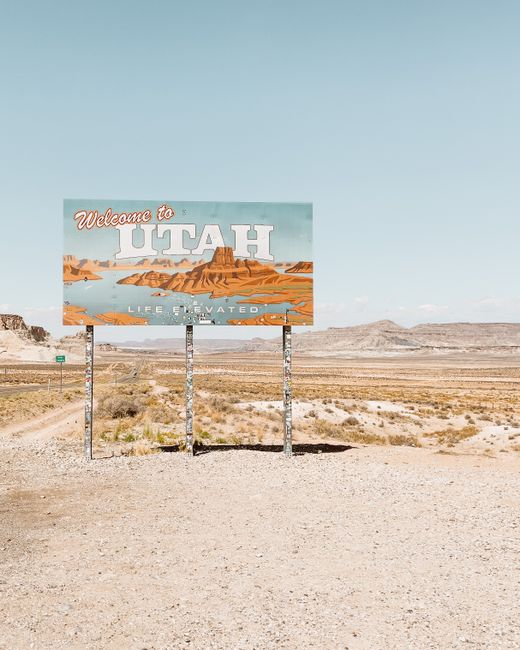 A 'Welcome to Utah' signboard stands tall in an arid desert landscape under a clear sky.