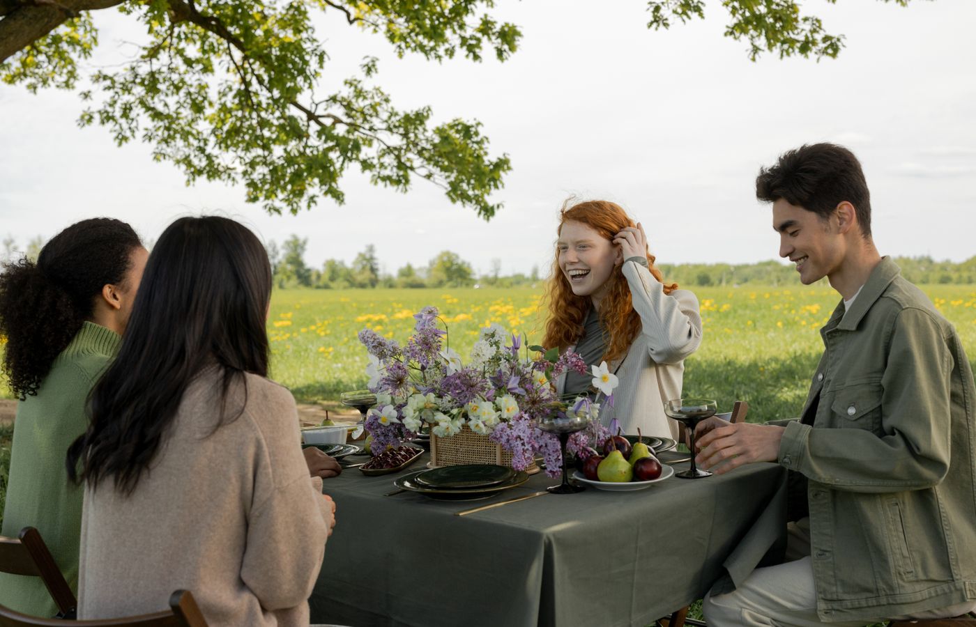 Group of friends enjoying a picnic in a meadow under a tree. Smiling and talking with drinks and a floral centerpiece.
