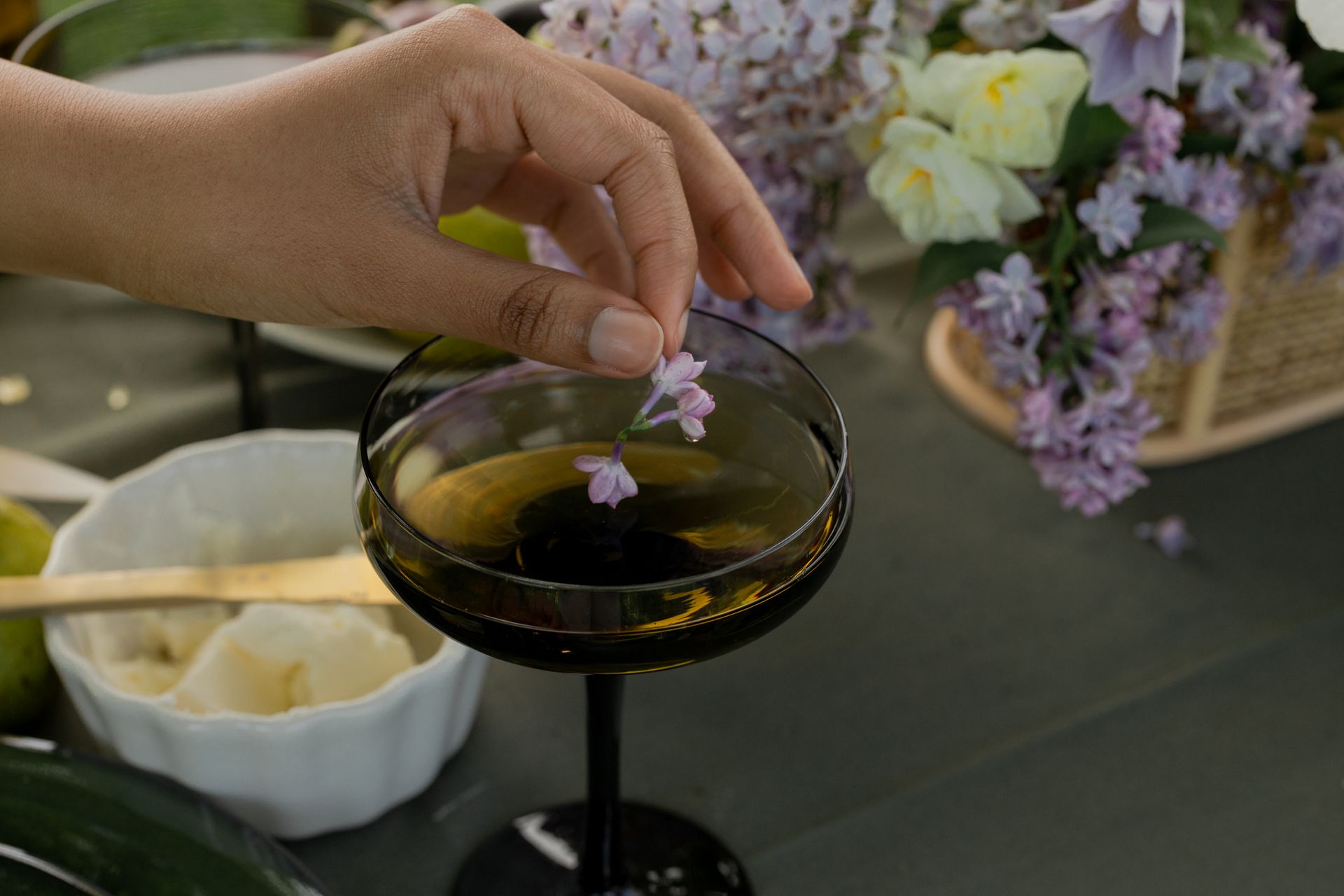 A hand gently places a lilac flower in a glass, next to a floral arrangement, outdoors.