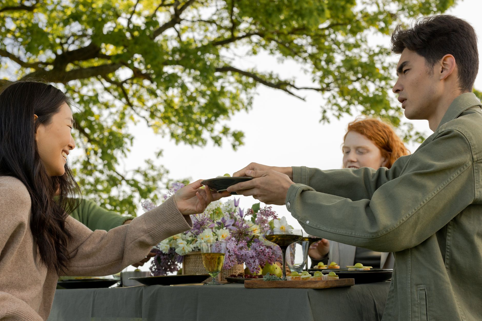A long wooden table set for a group meal outdoors under trees, plates and bowls laid out