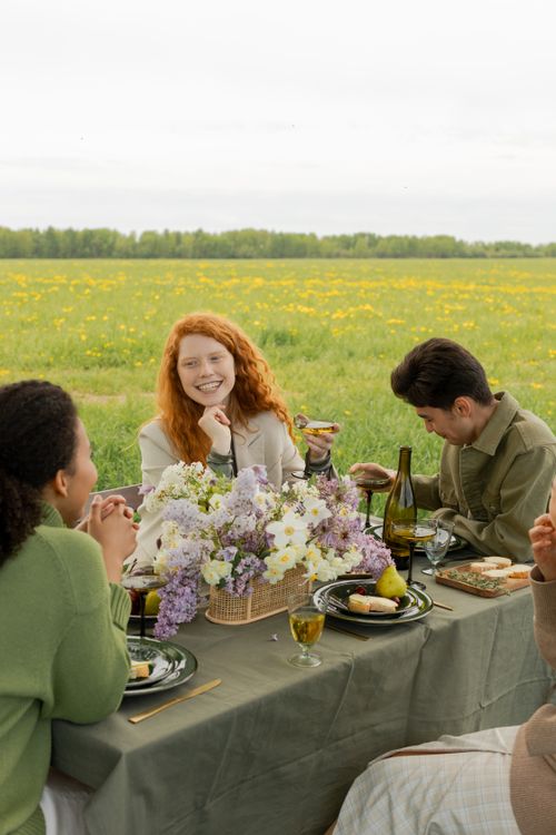 Free Friends laughing and enjoying a picnic with wine in a flower-filled field. Stock Photo