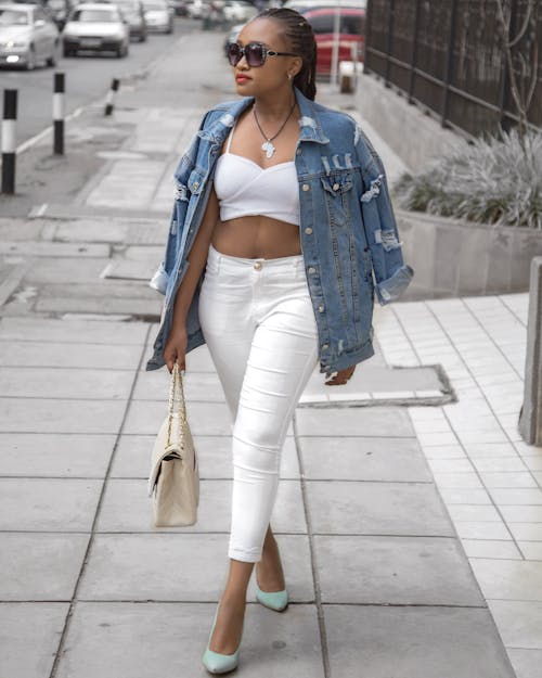 Free Photo of Woman Wearing White Crop Top and Denim Jacket Walking in Pavement Holding Bag Stock Photo