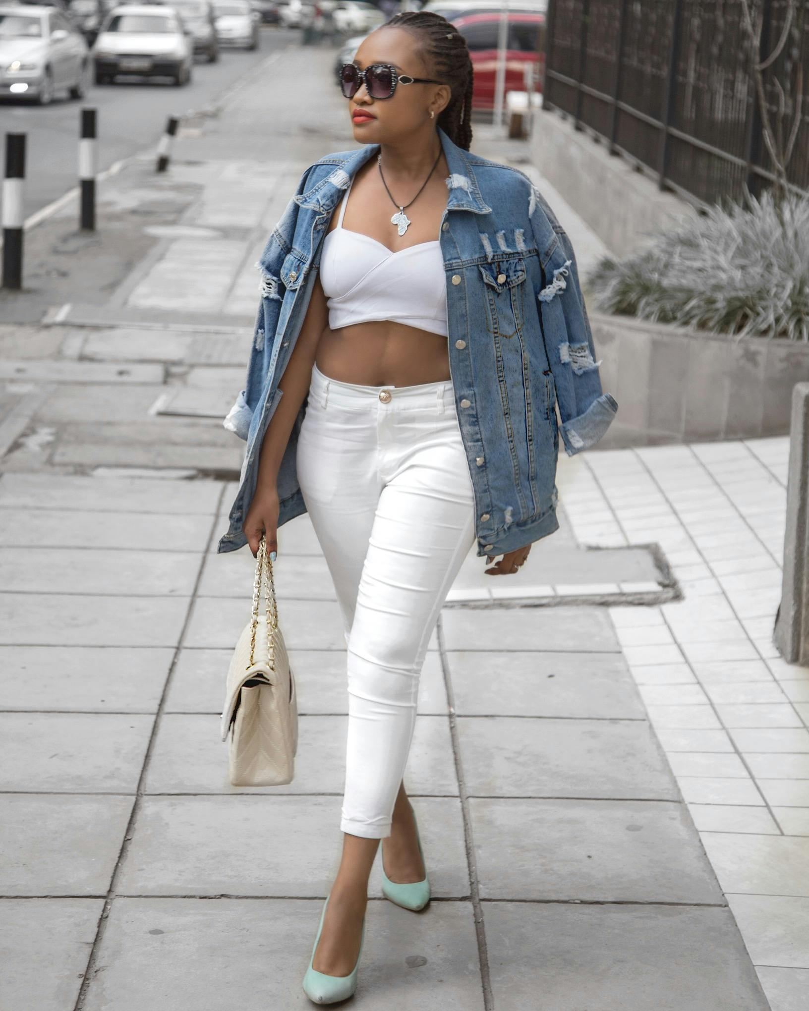 Photo of Woman Wearing White Crop Top and Denim Jacket Walking in Pavement Holding Bag