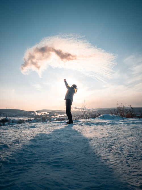 Free A silhouette of a person in winter clothing raising arms with backlit snowflakes under a bright sky. Stock Photo