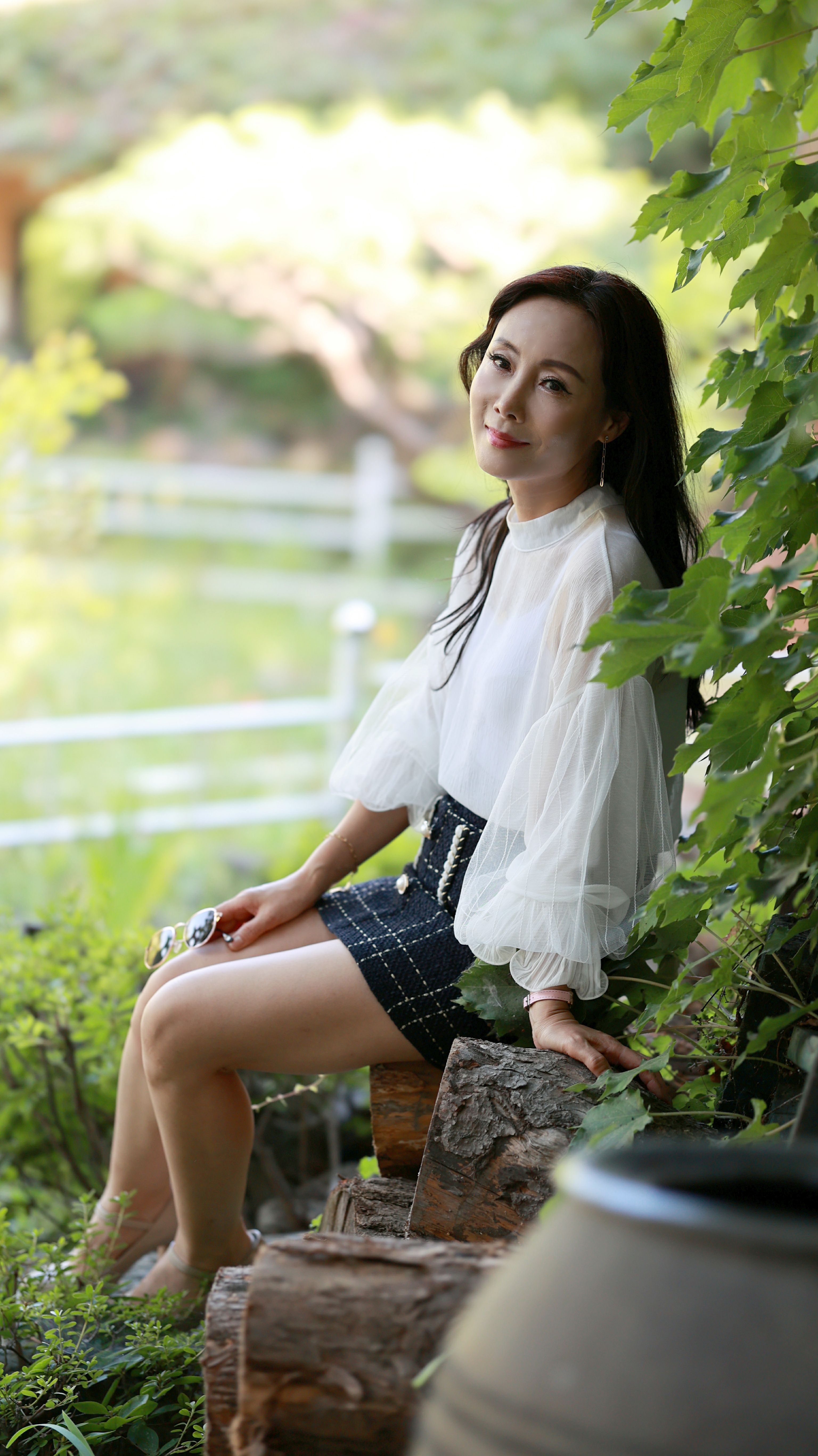 A serene portrait of a woman in a garden, wearing a white blouse and smiling at the camera.