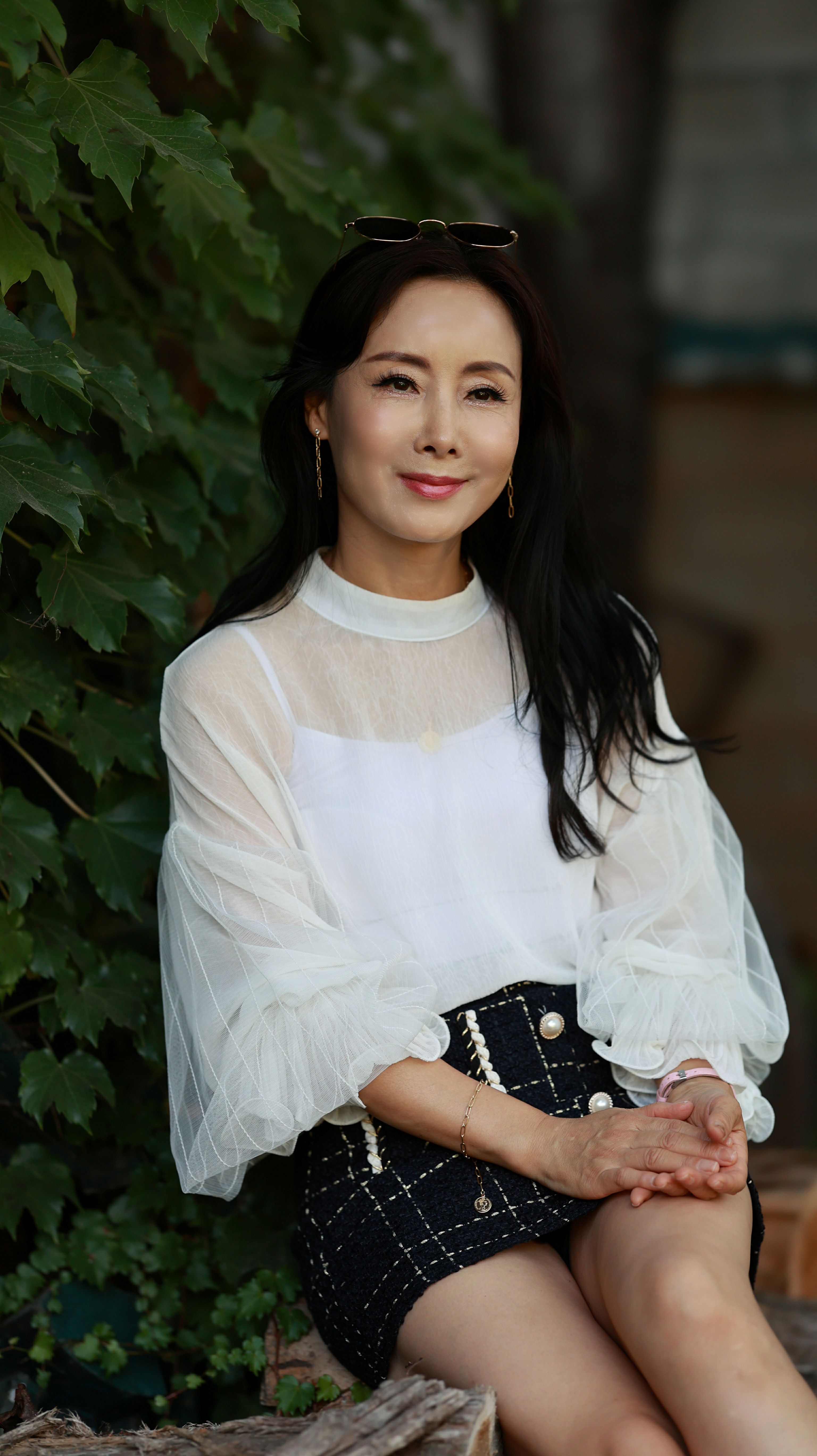 Portrait of a woman sitting outdoors in a white top and black skirt, surrounded by green leaves.