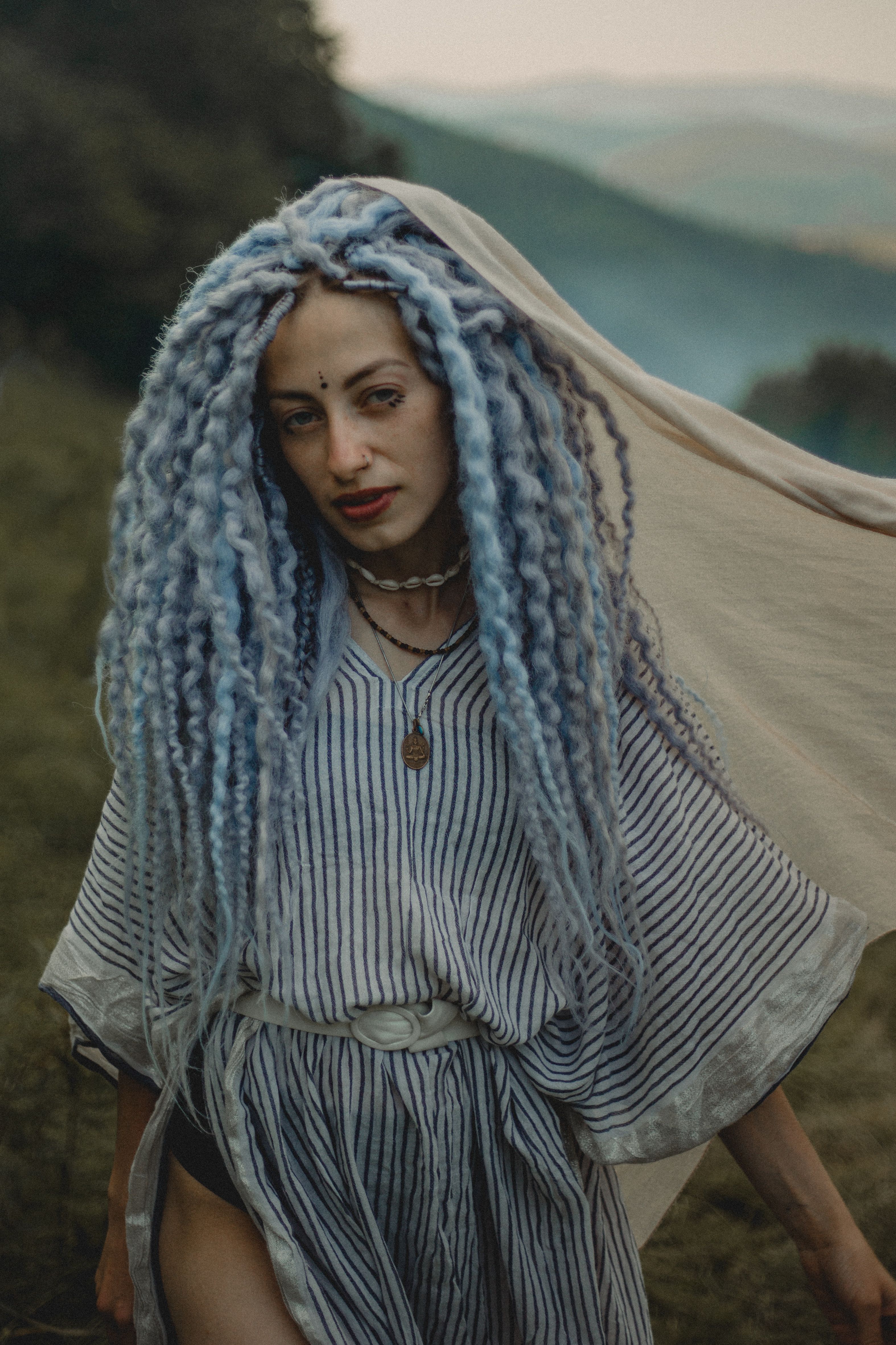 A woman with blue dreadlocks wearing a striped dress poses outdoors, exuding bohemian style.