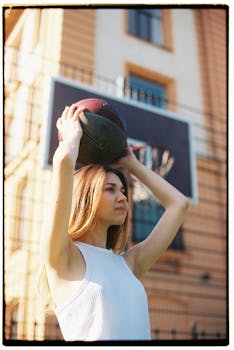 A young woman holds a basketball above her head outdoors, ready to throw at sunset.