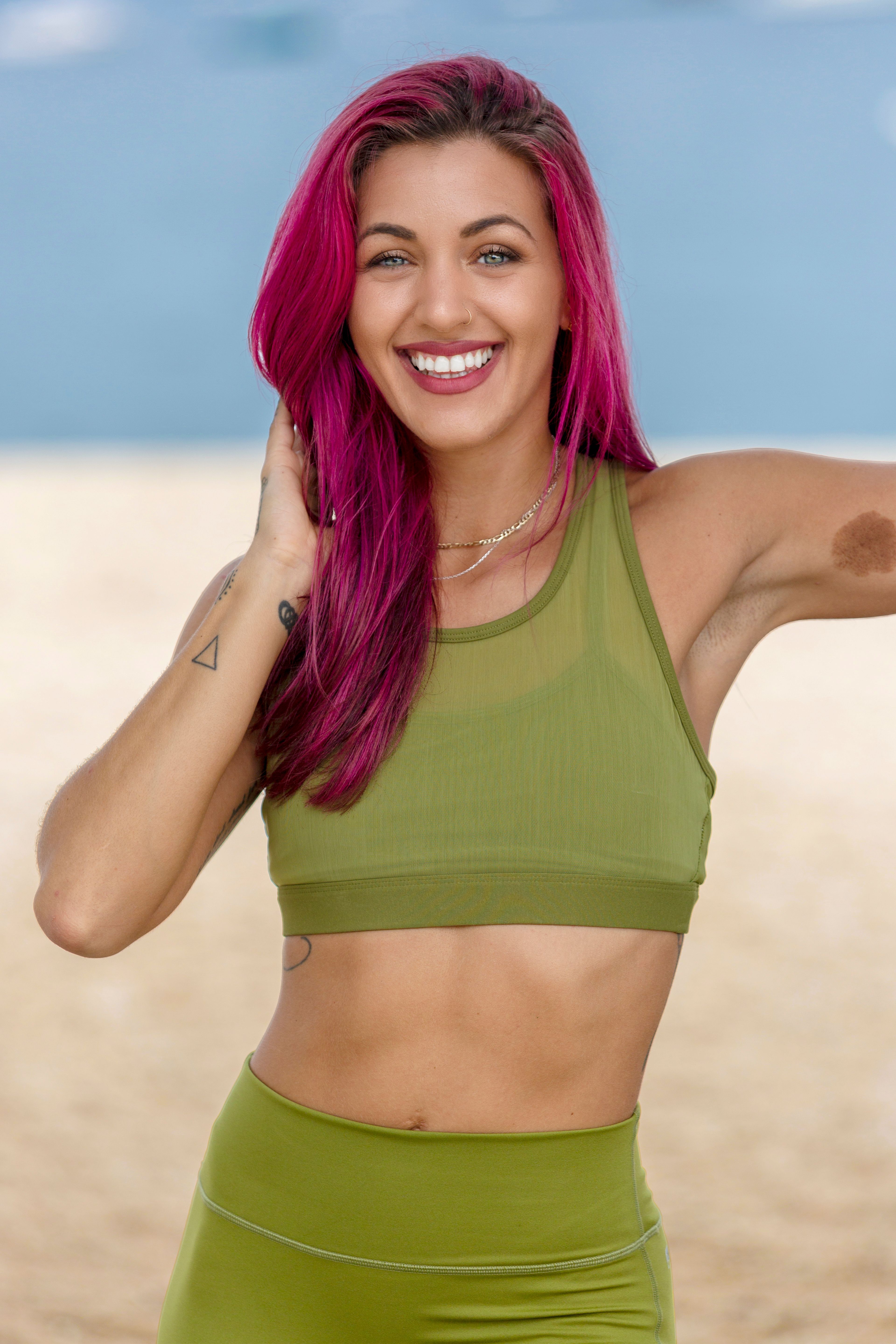 A smiling woman with vibrant pink hair in green sportswear posing confidently outdoors.