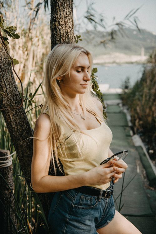 Free A blond woman leaning against a tree, enjoying a serene outdoor summer day by the water. Stock Photo