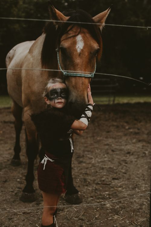Free A child in a costume with face paint lovingly pets a horse in a rustic outdoor setting. Stock Photo