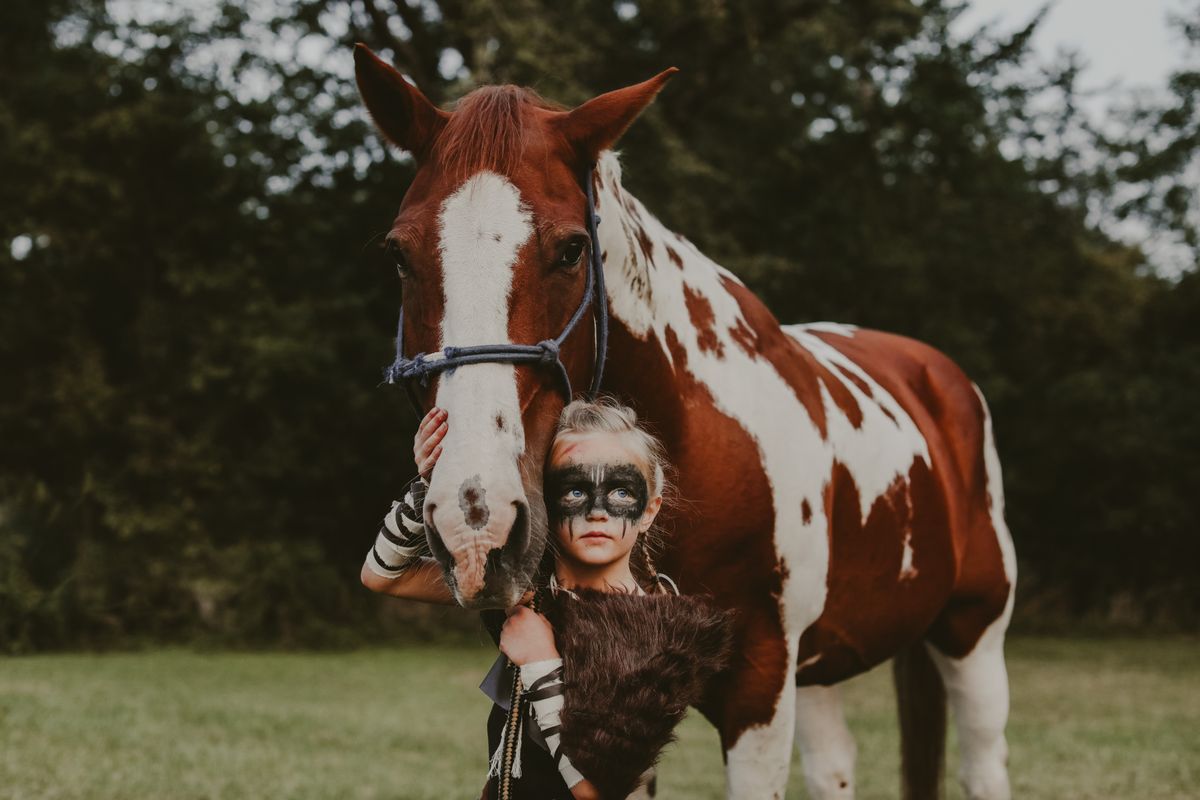 A child with playful face paint standing beside a brown and white horse