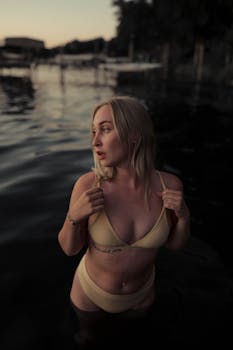 A woman in a bikini stands in calm waters during twilight, surrounded by a peaceful lakeside setting.
