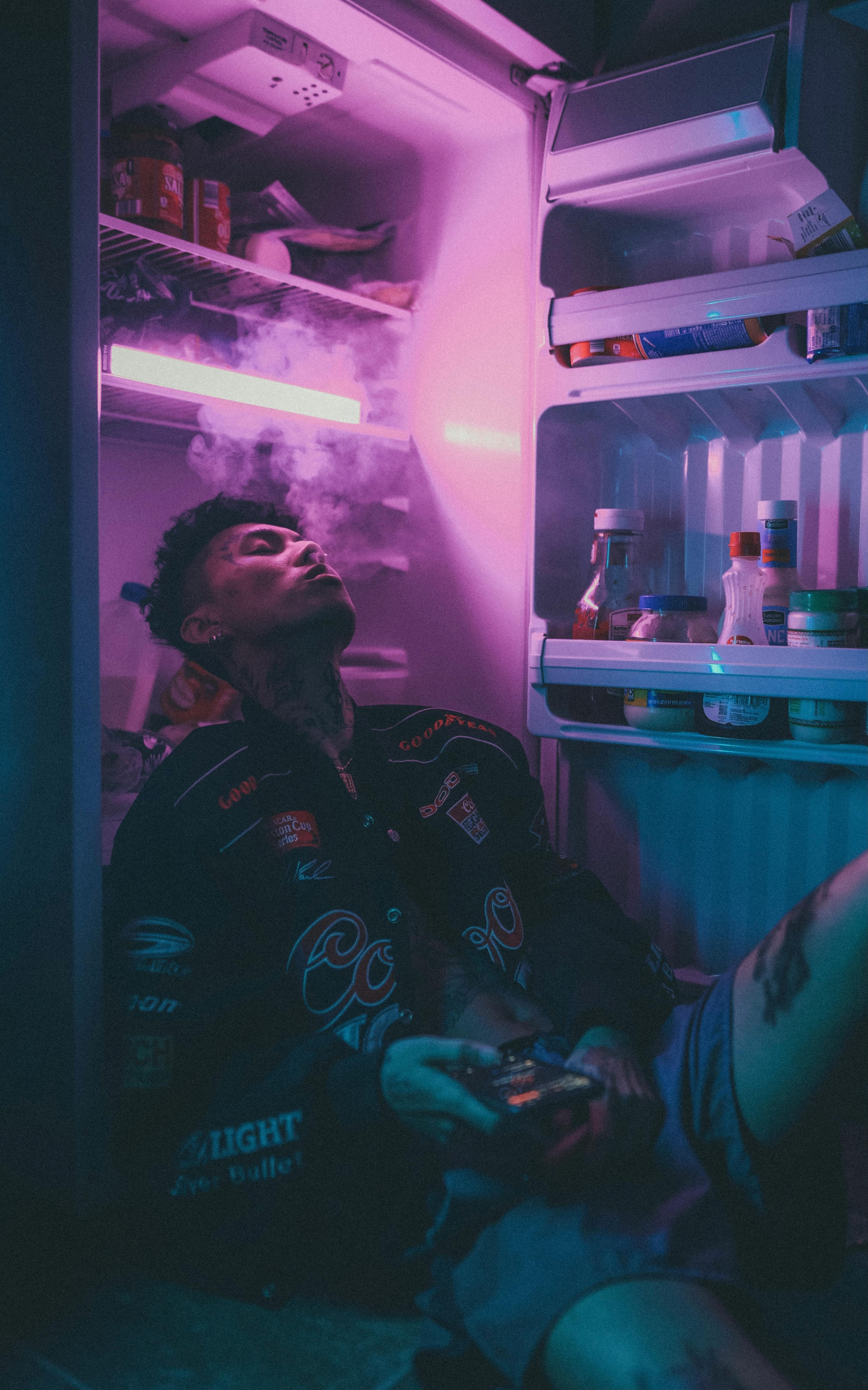 A person seated in front of an illuminated refrigerator emitting vibrant neon light inside.