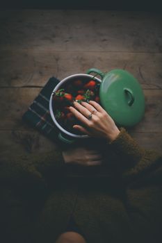 A hand touches fresh strawberries in a green pot with a moody flat lay composition.