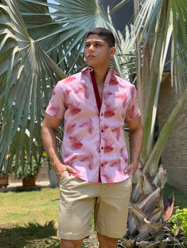 A man in a vibrant tropical shirt stands by a tall palm tree in a sunny garden setting.