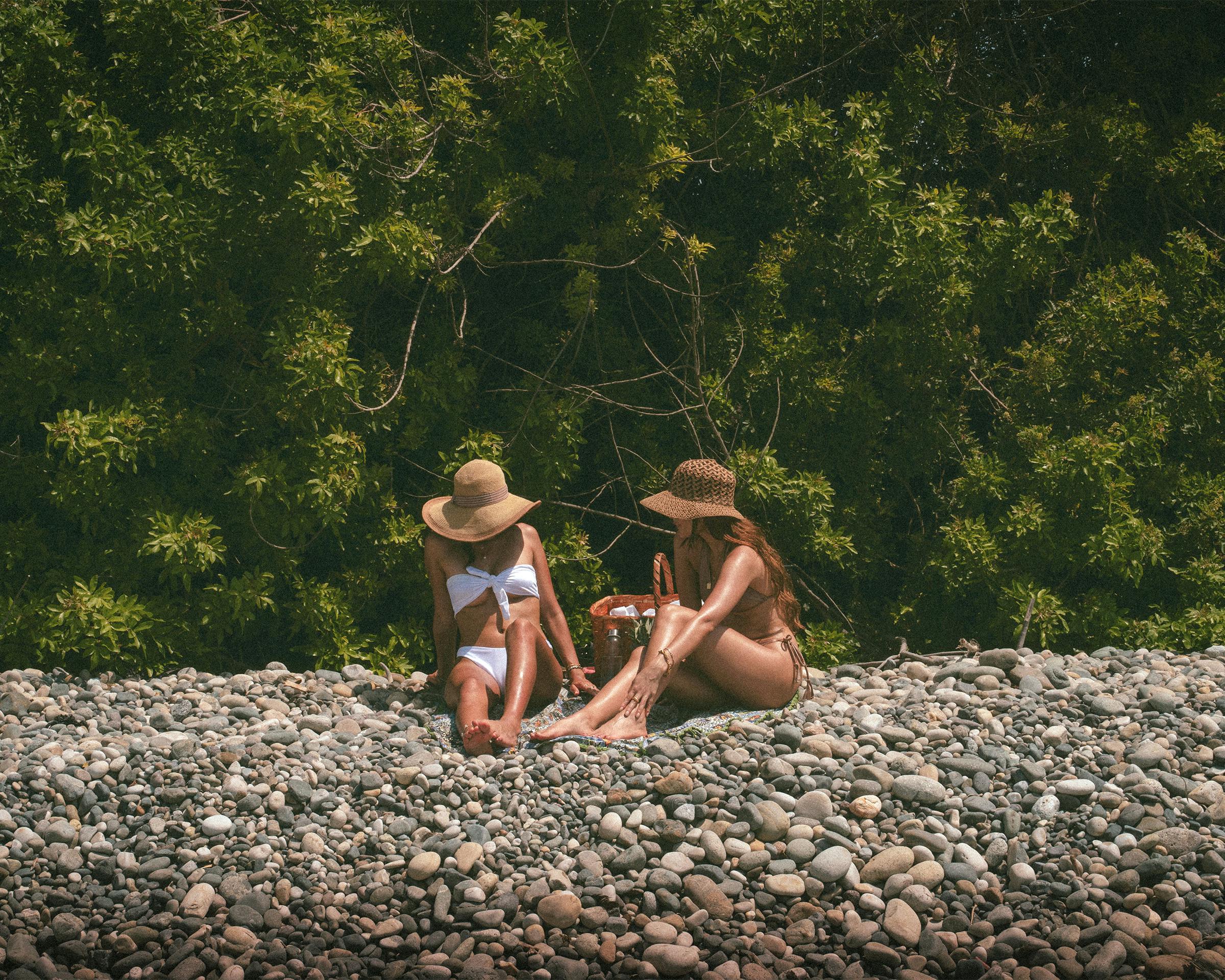 Two women in swimsuits enjoying a sunny beach day, surrounded by pebbles and greenery.