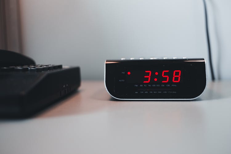 Digital Alarm Clock Beside A Telephone On A White Surface