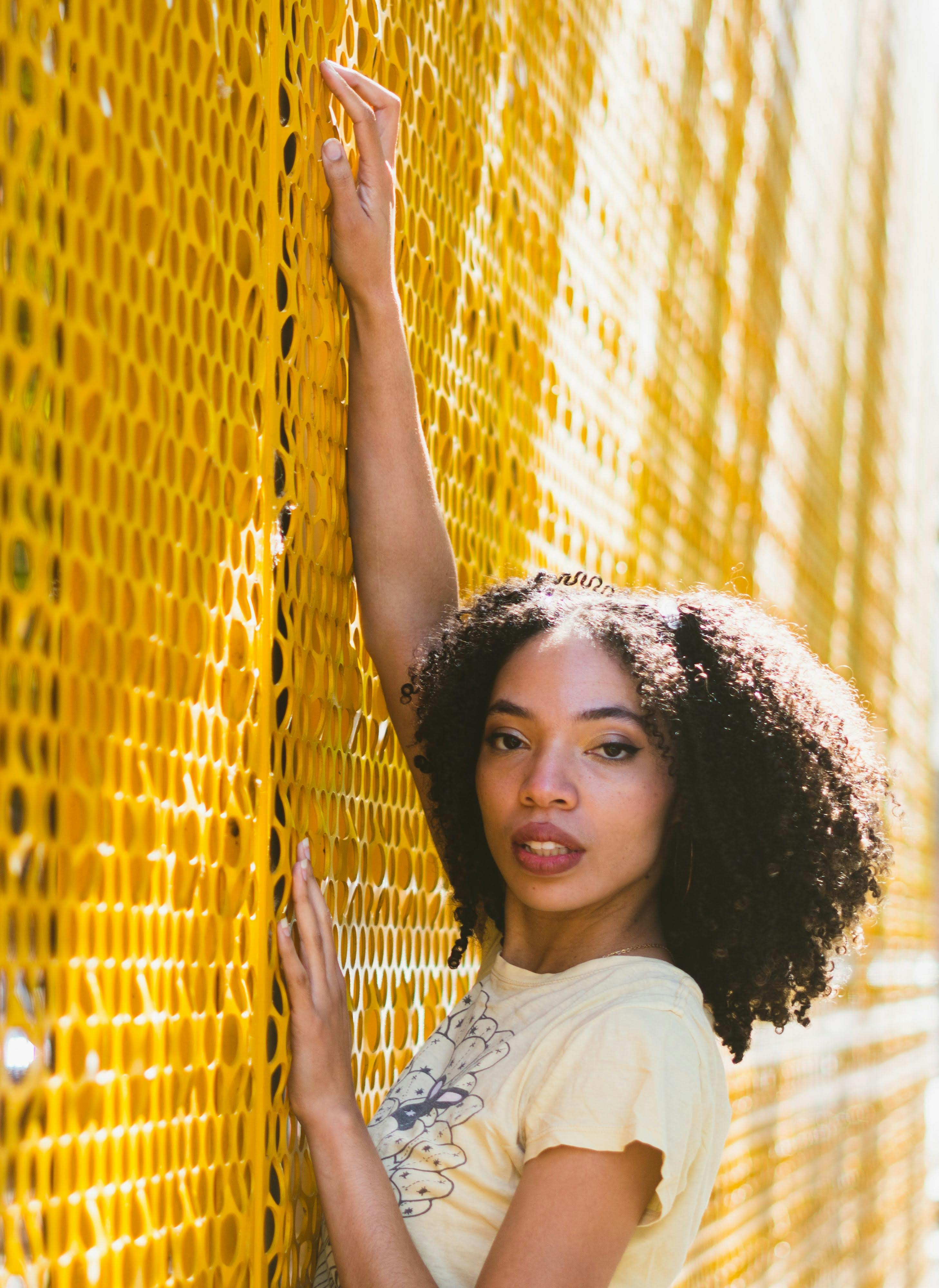 Stylish woman with afro hair poses confidently against a vibrant yellow mesh fence outdoors.