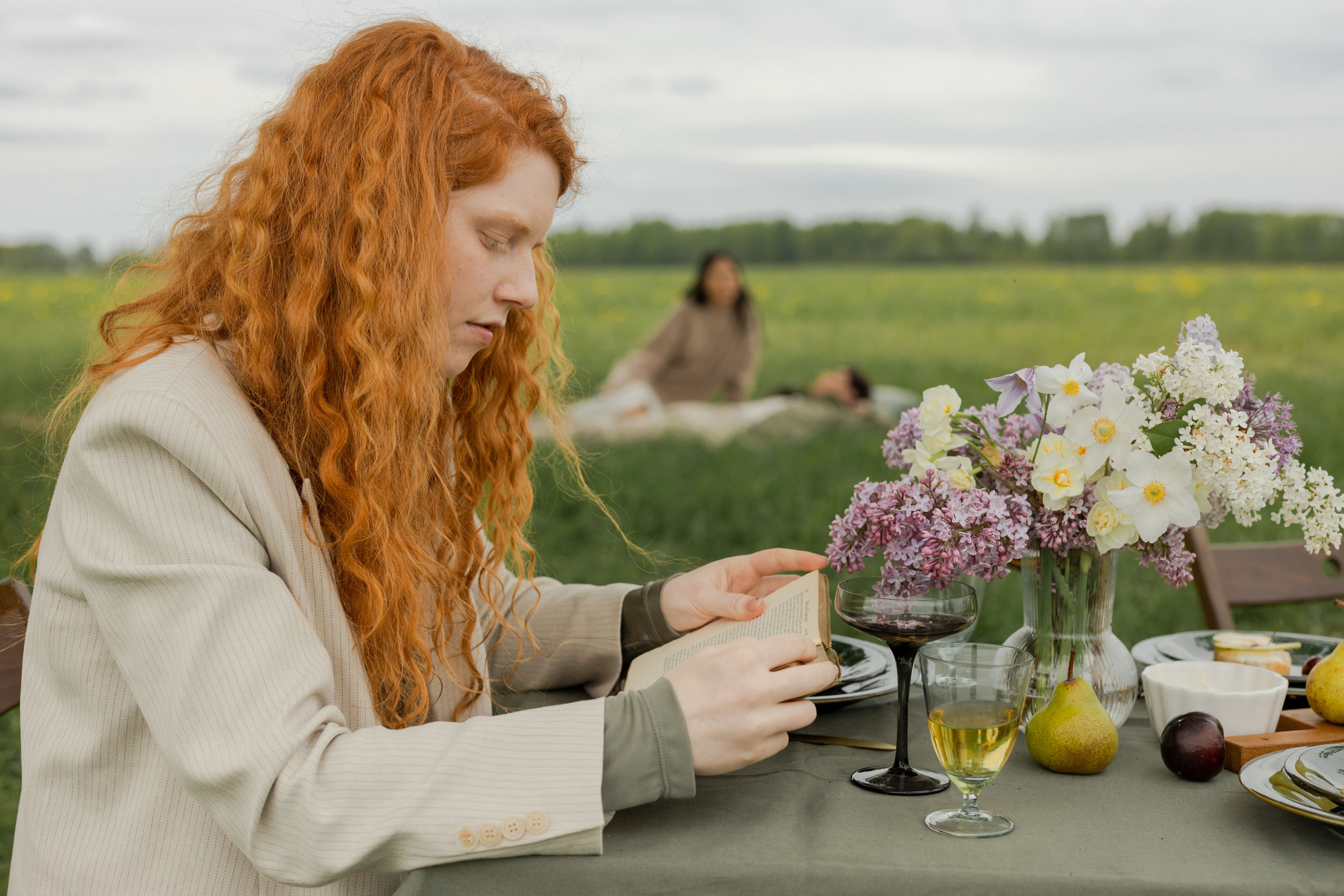 A tranquil outdoor picnic scene with a woman reading among flowers and drinks.