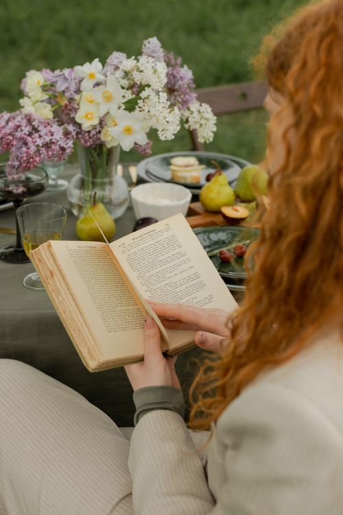 Free A woman enjoying a book at a beautiful outdoor picnic with flowers and fruits. Stock Photo