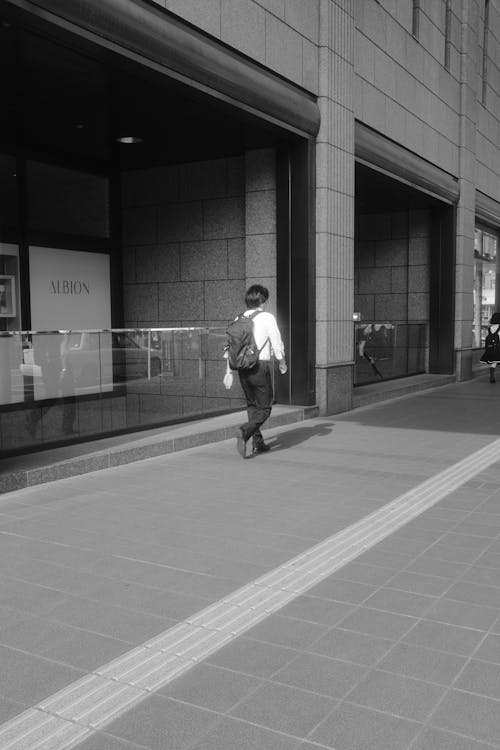 Grayscale Photo of a Person Walking on a Sidewalk