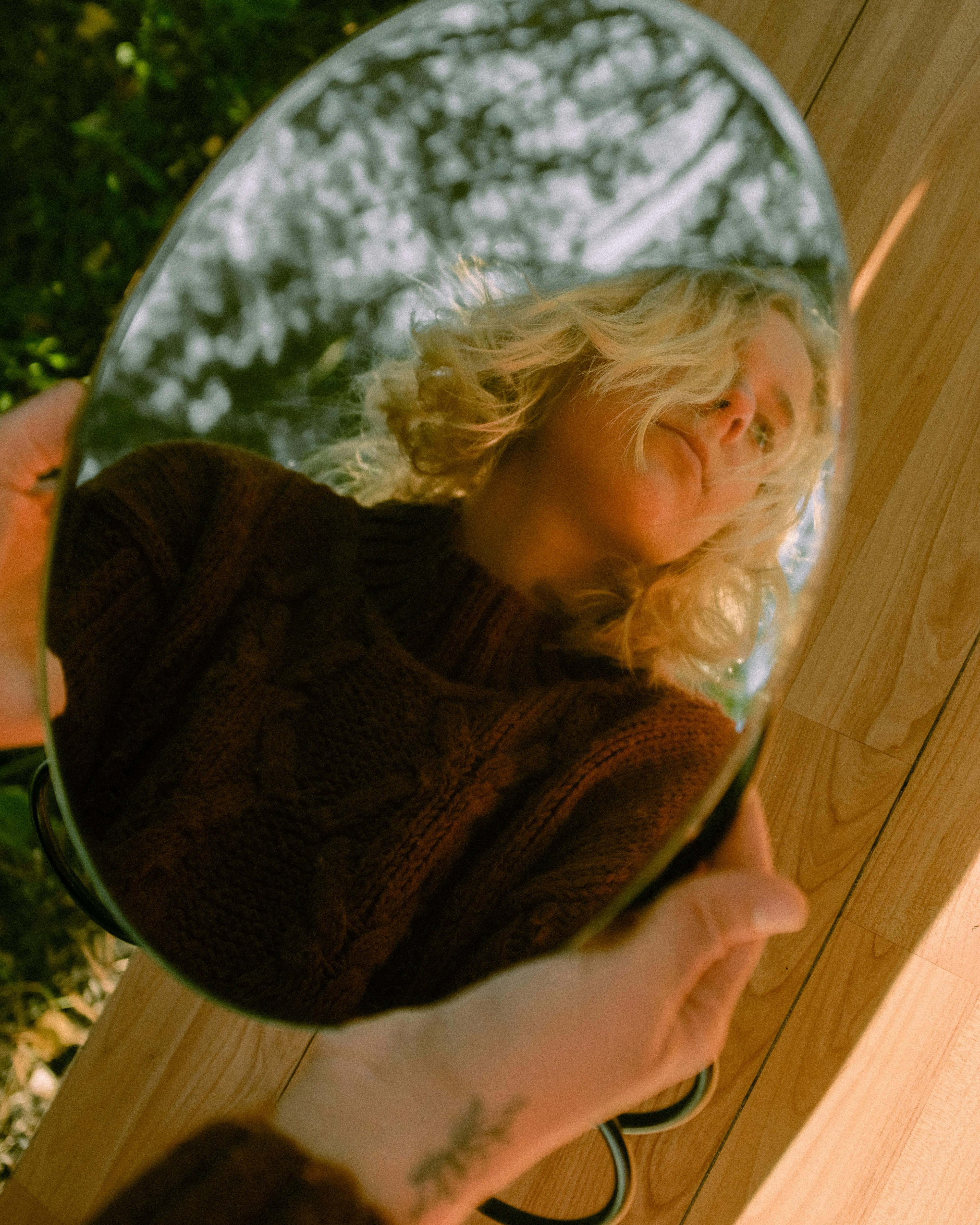A woman in a sweater reflecting in a handheld mirror outdoors on a sunny day.