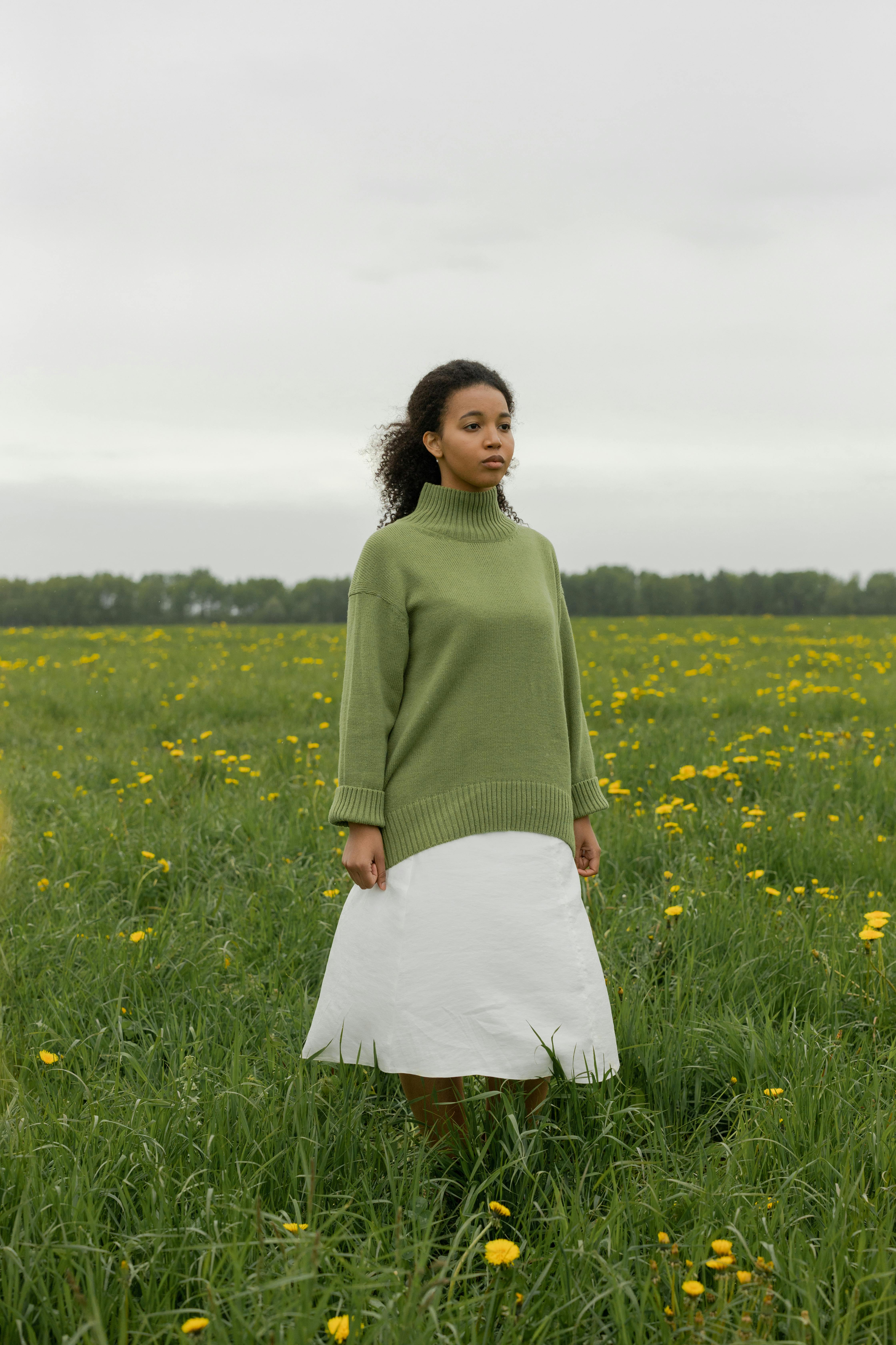 A young woman stands in a meadow wearing a green sweater and white skirt, surrounded by blooming flowers.