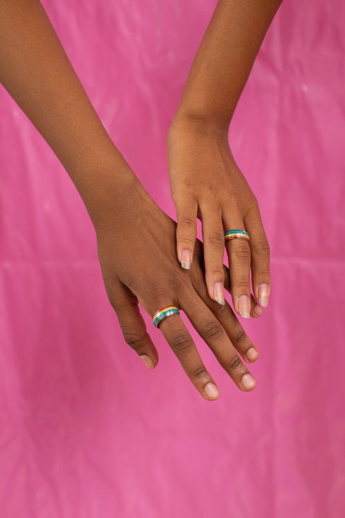 Free A close-up of two diverse hands wearing rainbow rings symbolizing LGBTQ pride on a pink background. Stock Photo