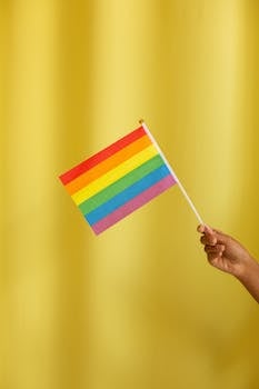 Hand holding a rainbow flag symbolizing LGBT pride against a vibrant yellow backdrop.