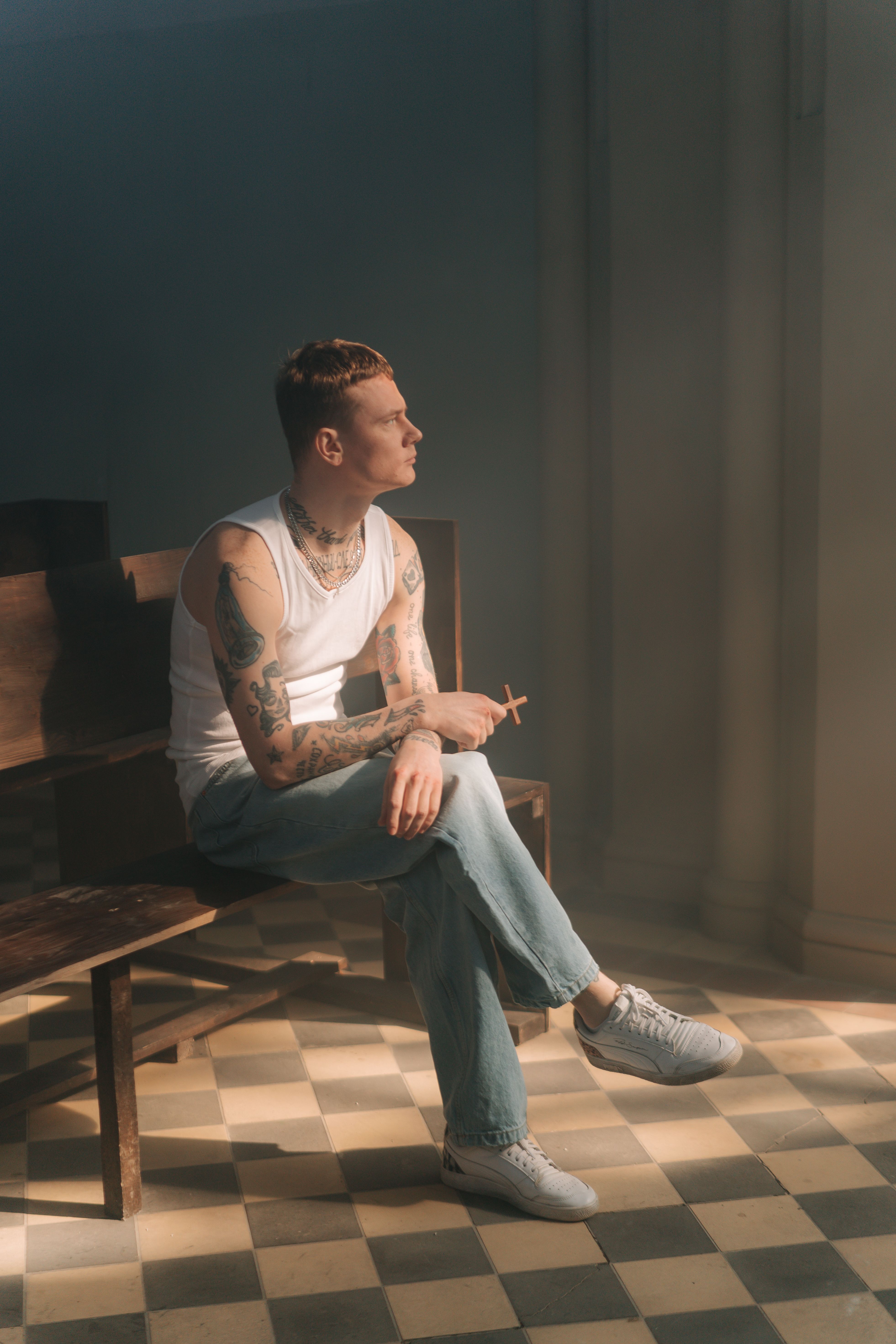 Tattooed man in tank top holding a cross, sitting in thoughtful contemplation on a pew.