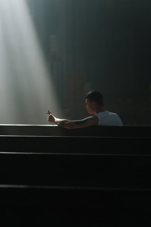A Man in White Tank Top Sitting on a Pew while Holding a Cross
