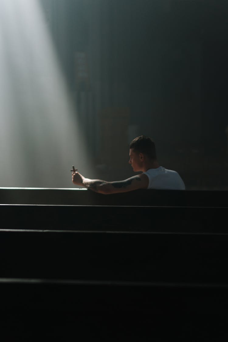 A Man In White Tank Top Sitting On A Pew While Holding A Cross